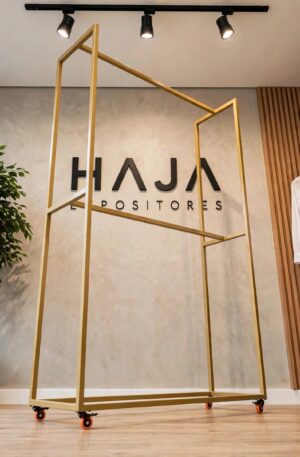 Arara Dourada de Roupa Dupla Alta com Rodas de 2.25 metros de altura por 1.5 metros de largura e 0.45 metros de profundidade, com pintura eletrostática fosca, posicionada em showroom elegante com o logotipo da HAJA Expositores ao fundo.